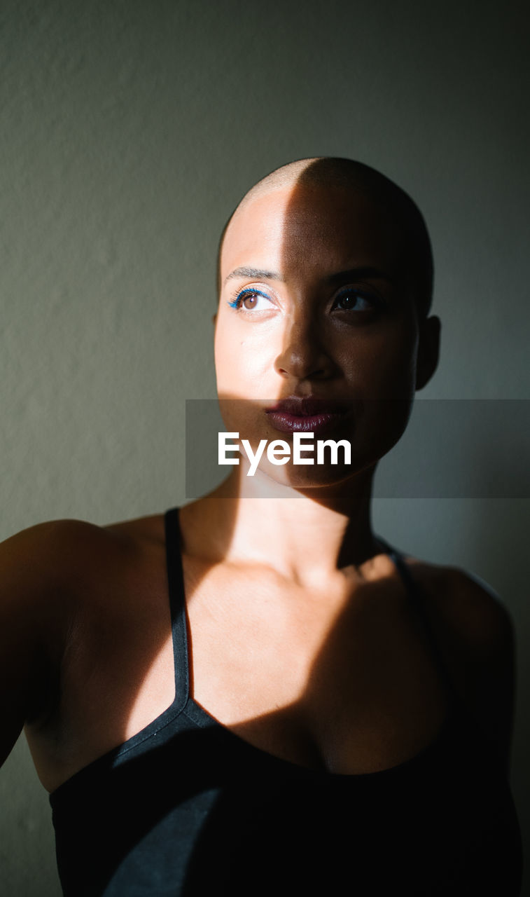 Thoughtful african american female with bald hairstyle standing near window in apartment and illuminated by sunbeam in morning