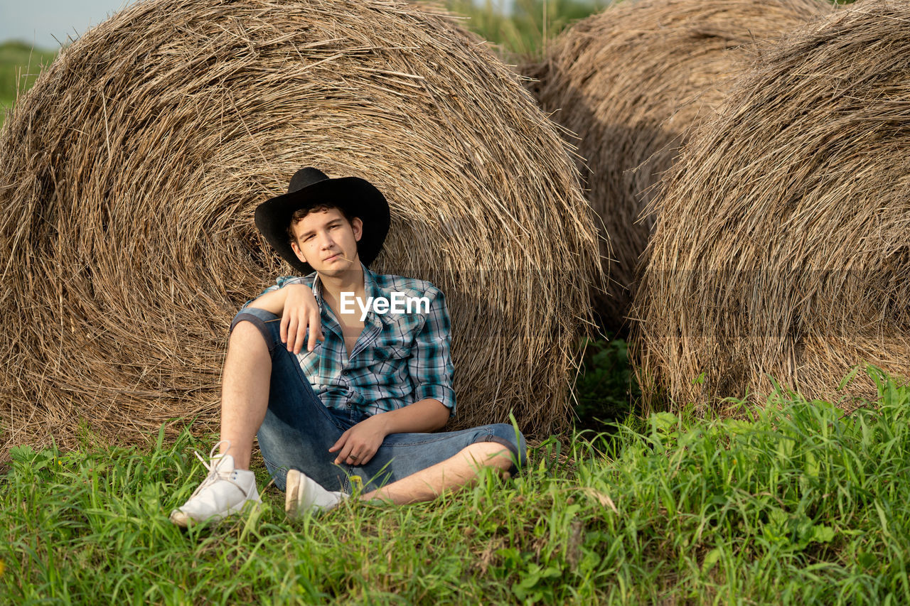 Full length of young man sitting on field near the haystack 