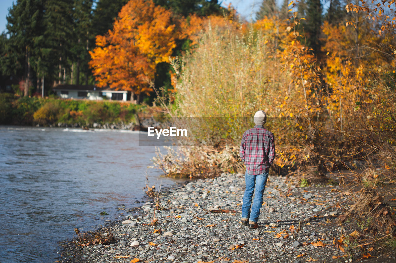 Teen boy in jeans, plaid shirt, and beanie walks along river in fall.