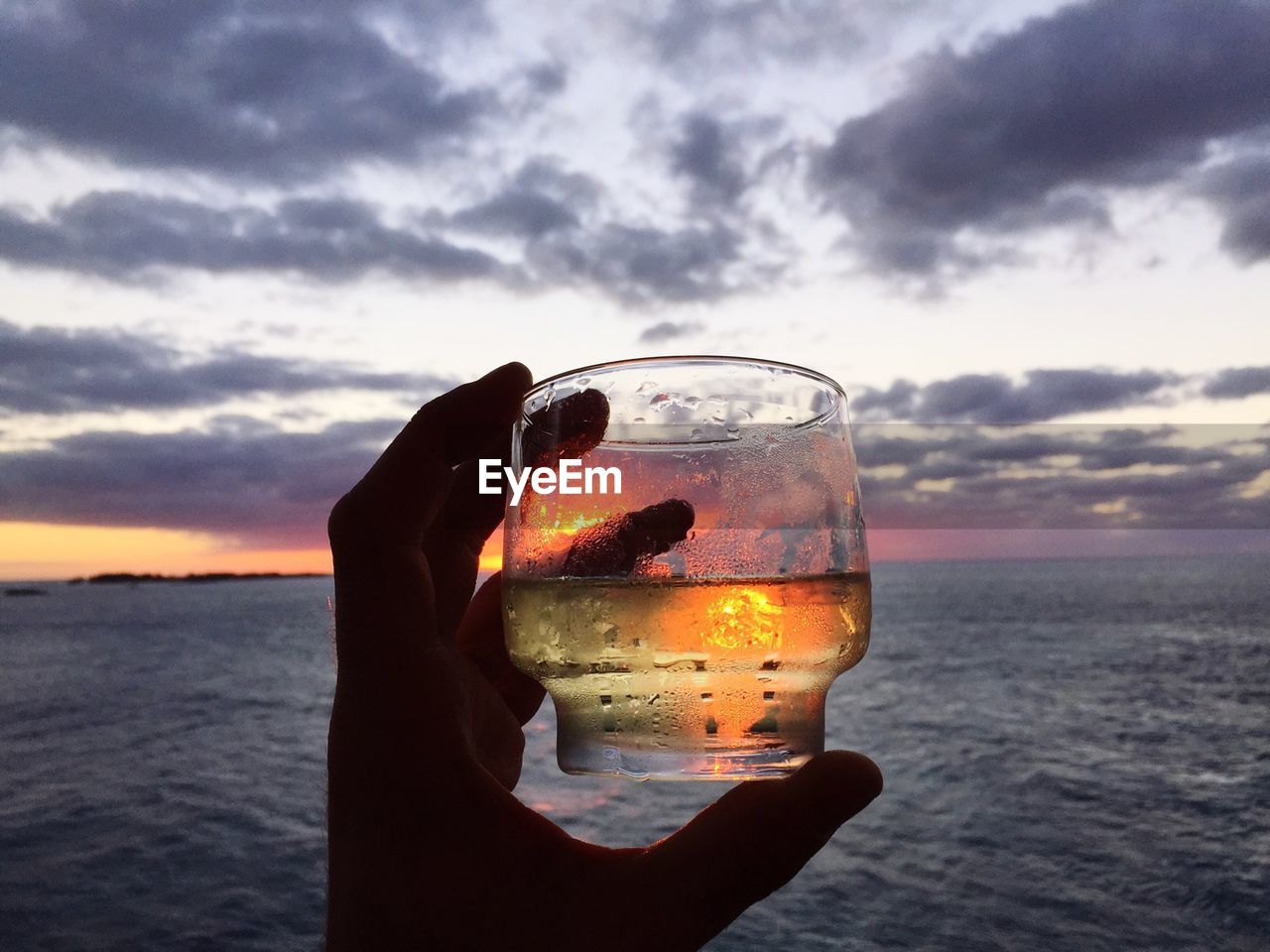 Cropped image of person holding drink at shore against cloudy sky