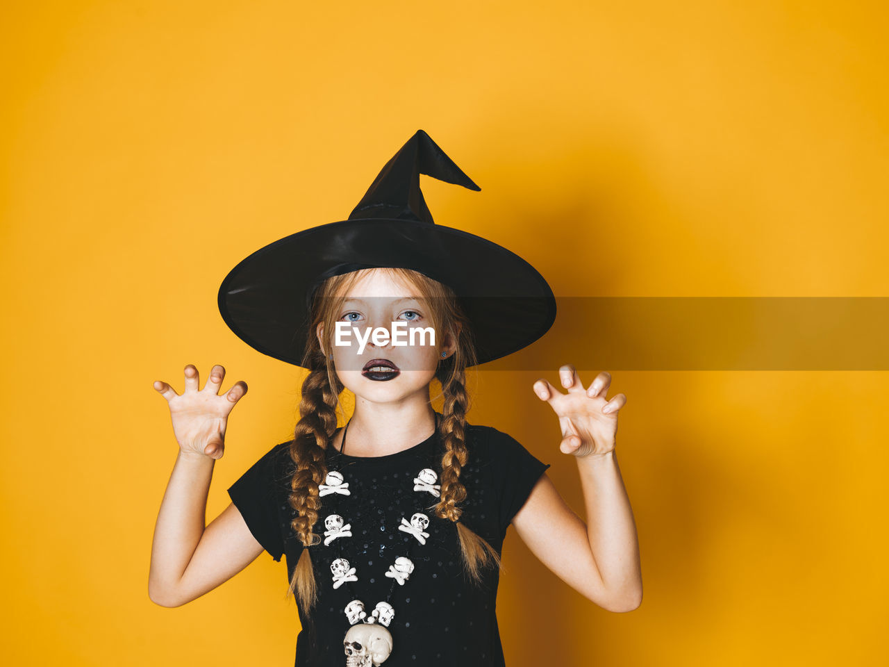 Portrait of girl wearing witch hat gesturing against yellow background during halloween