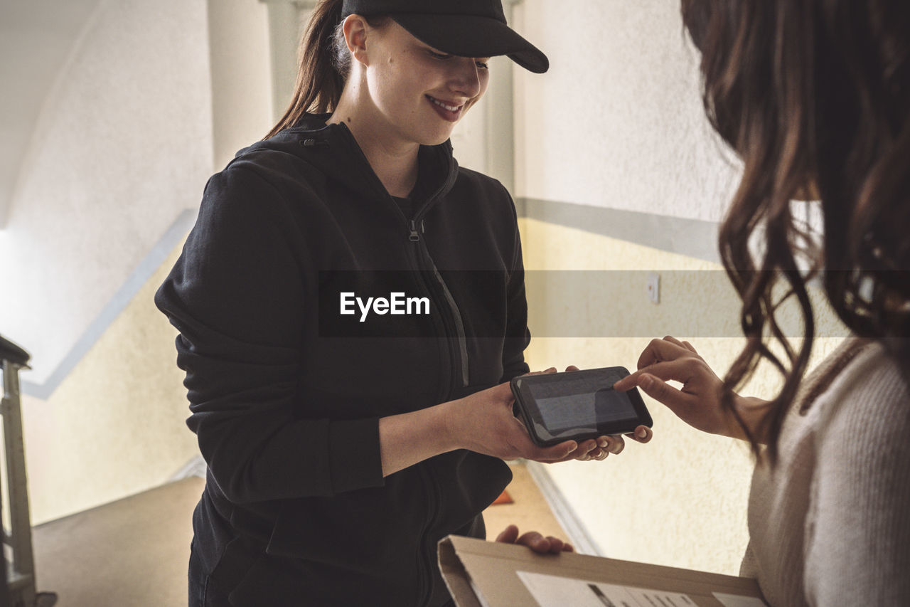 Smiling delivery woman holding phone while customer signing during home delivery