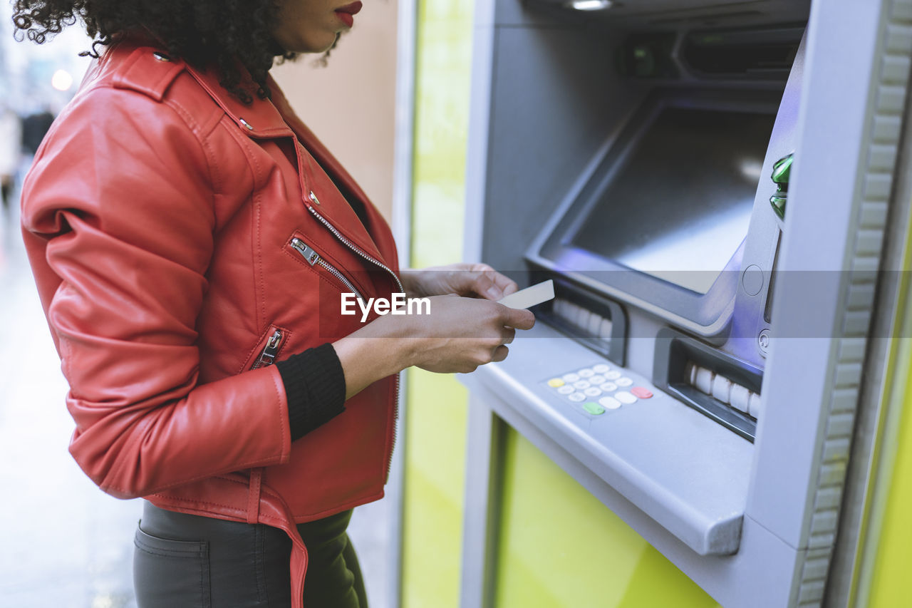 Anonymous african american female standing near atm and inserting credit card while smiling widely and looking away