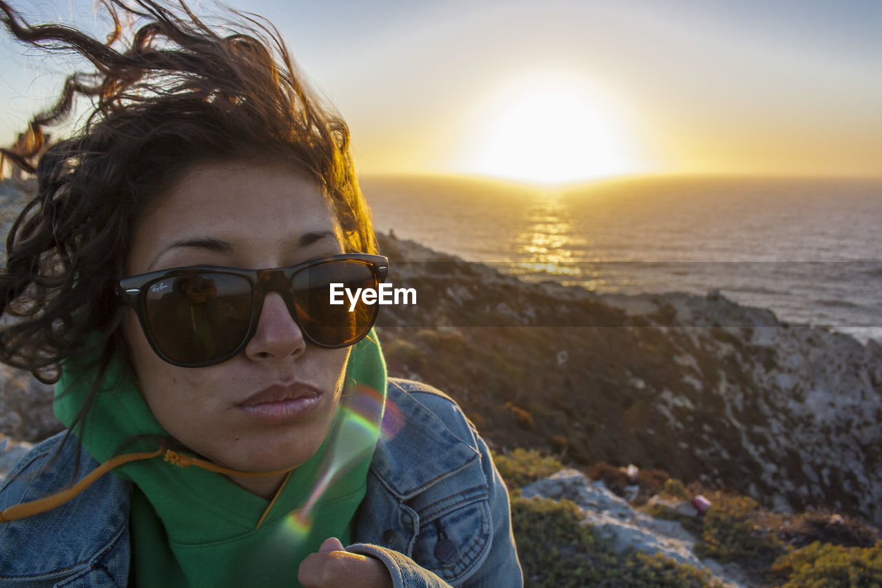 Young woman wearing sunglasses standing on cliff by sea during sunset