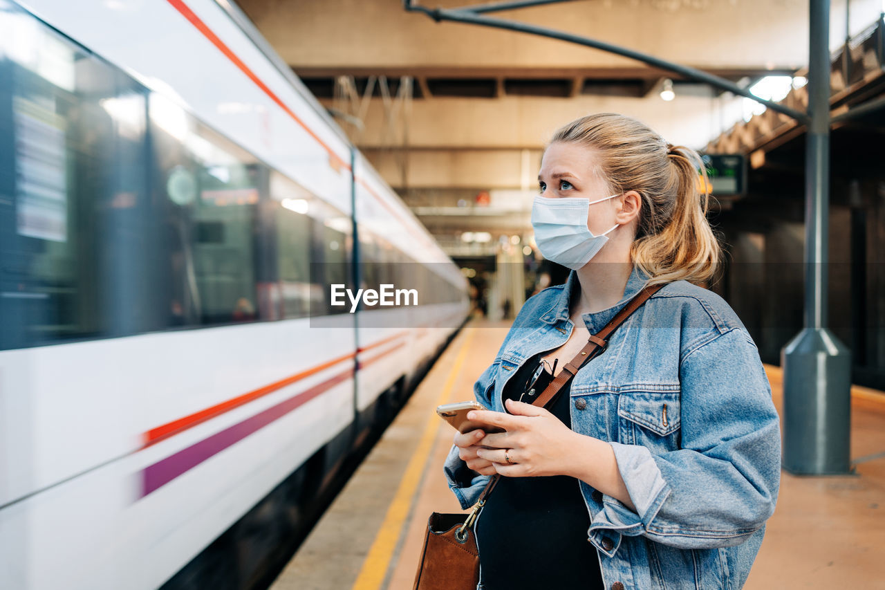 Young female in casual clothes and protective mask standing on railway station and looking away while waiting for train