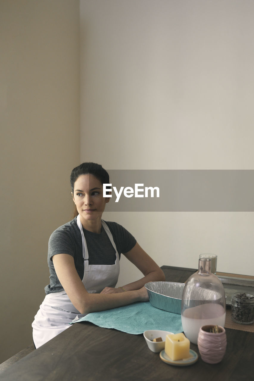 Thoughtful woman in apron sitting with containers and ingredients for preparing food at home