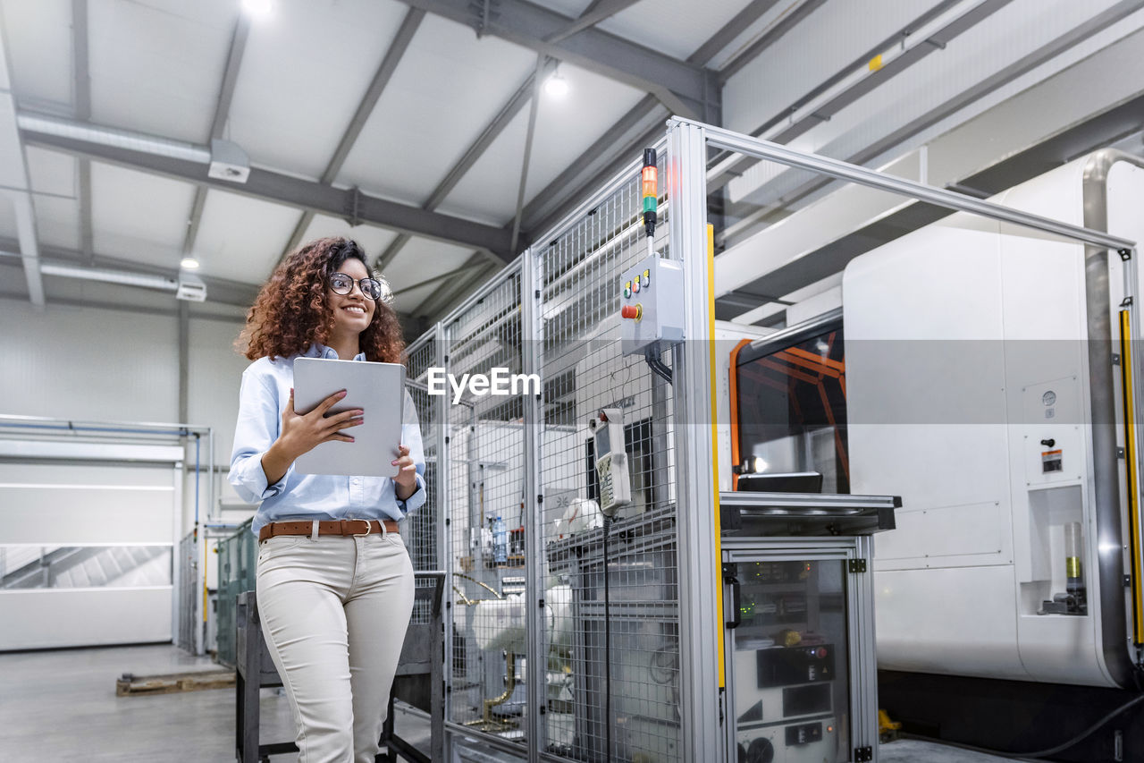 Smiling young engineer standing with tablet pc by machine in industry