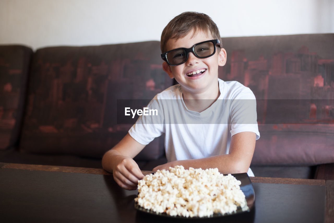 A boy in 3d glasses watches a movie and eats popcorn with his mouth wide open sitting on the couch