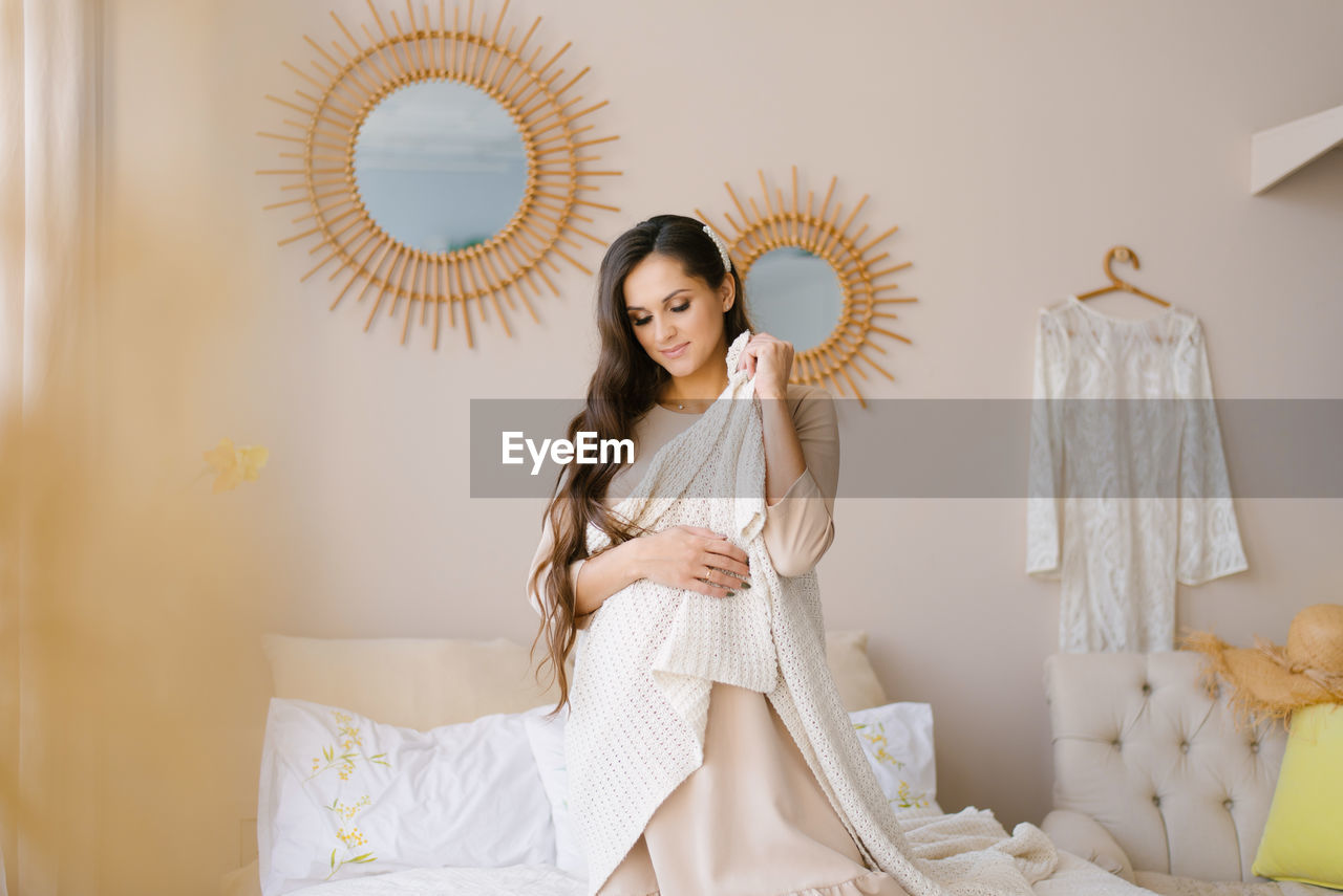 A pregnant young woman has thrown a blanket over her shoulders and is sitting on the bed waiting 