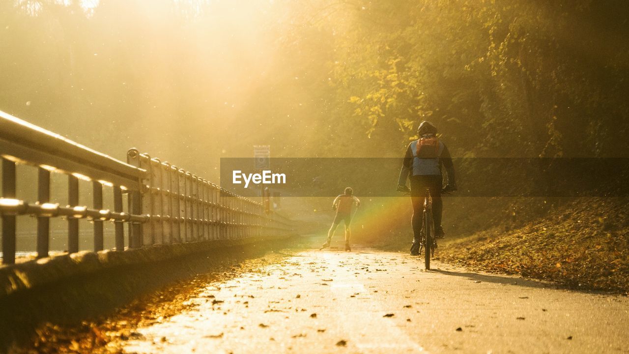 Rear view of man skating with friend riding bicycle on road during sunny day