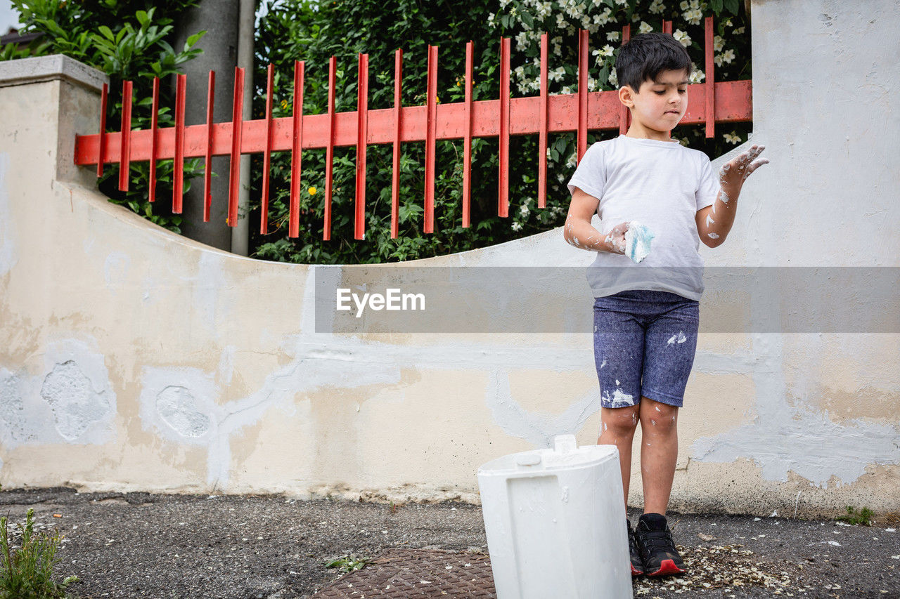 Standing child holding brush in one hand used for painting house fence wall