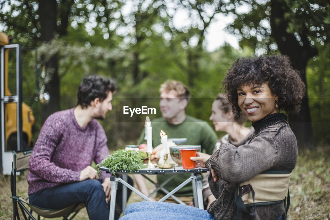 Portrait of smiling afro woman sitting with friends at table during camping in forest