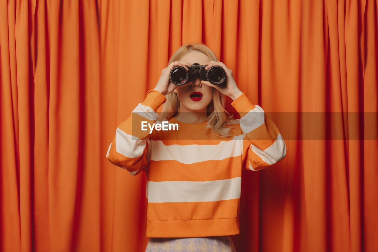 Woman looking through binoculars standing in front of orange curtain