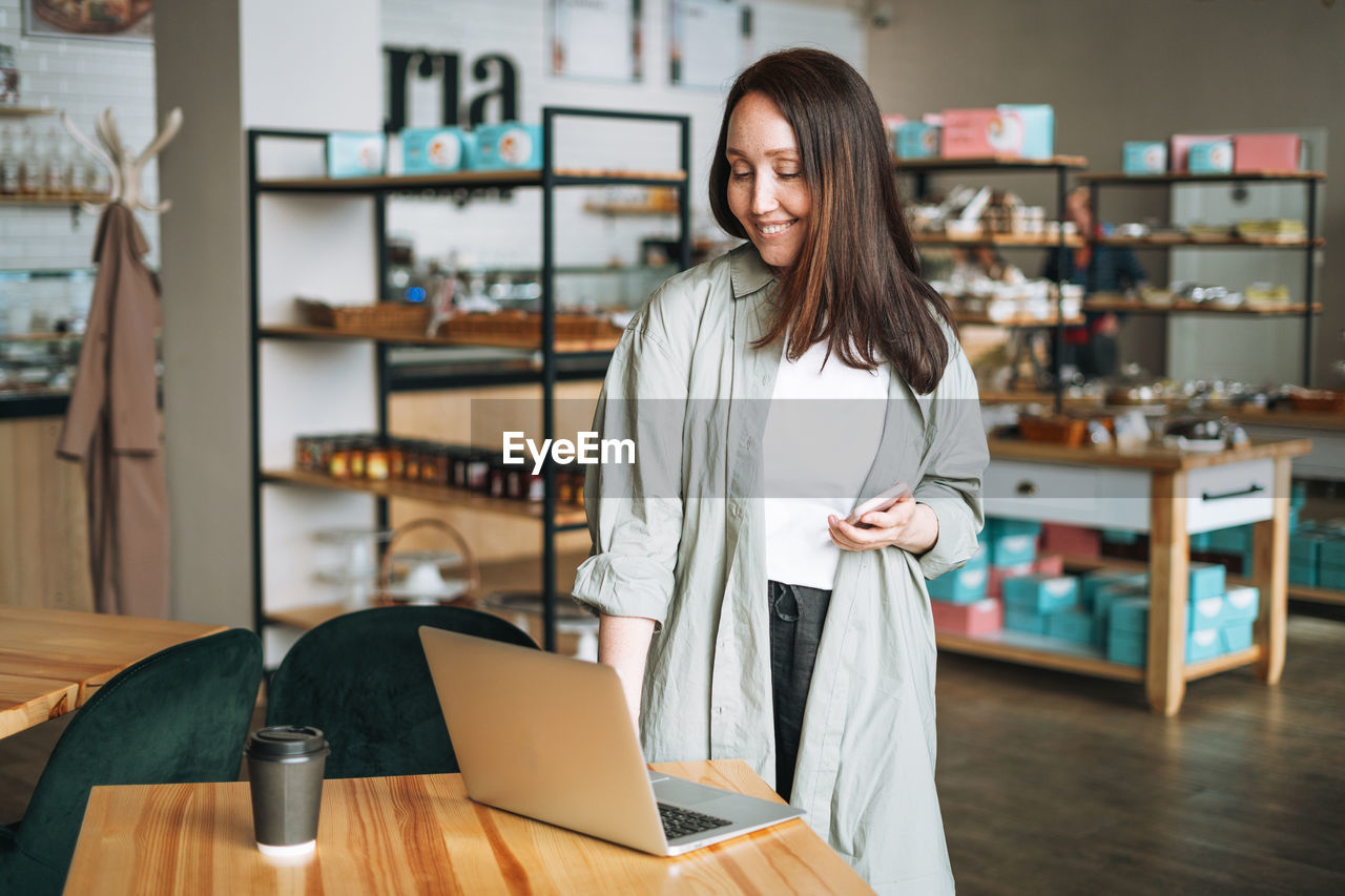 Adult business woman with long hair in stylish shirt working on laptop using mobile phone in cafe