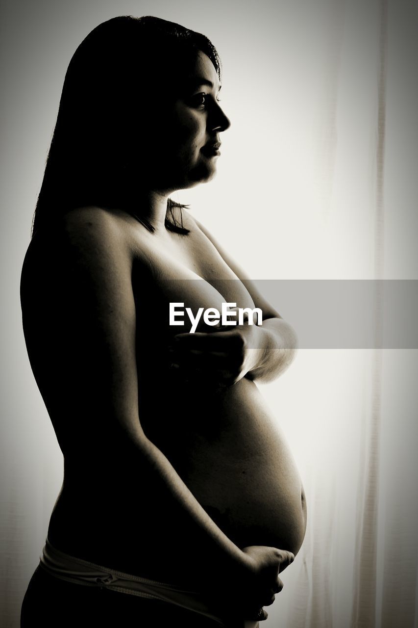 Shirtless pregnant woman with hands on stomach and breast looking away at home
