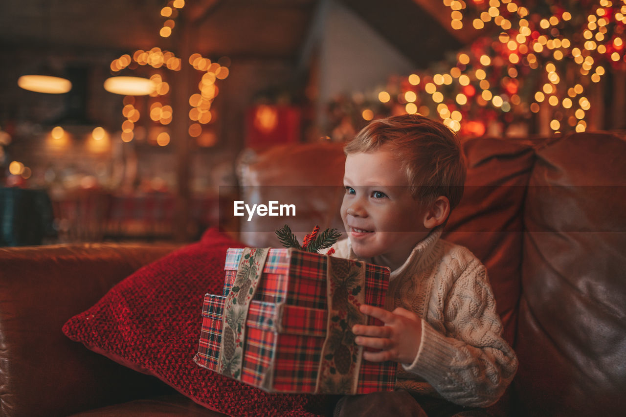 Candid authentic happy child in knitted beige sweater sitting with presents at lodge xmas decorated