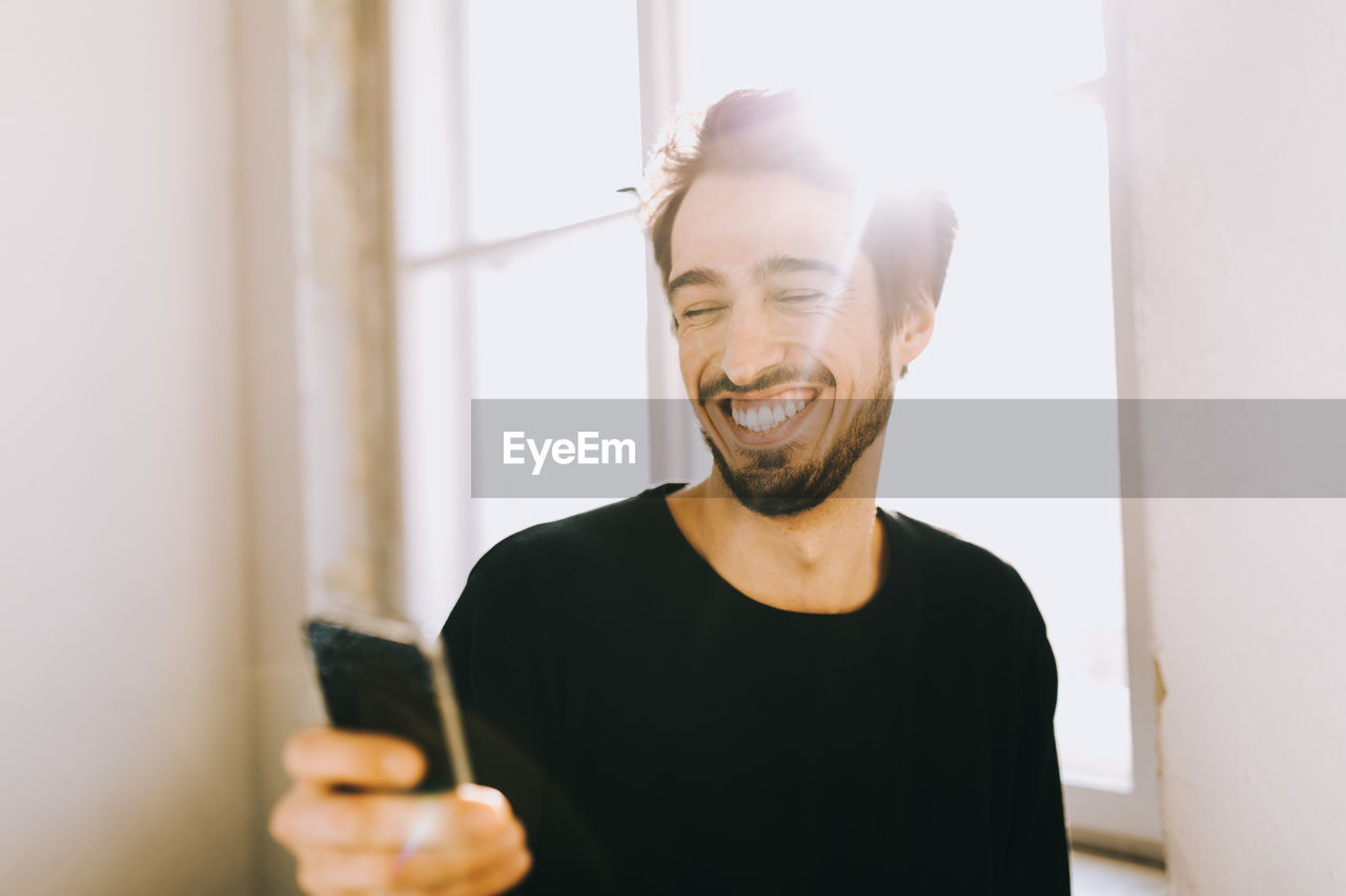 Happy young man using phone by window at home