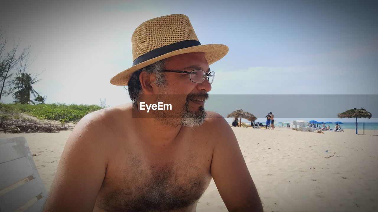 Shirtless man wearing hat and eyeglasses at beach against sky