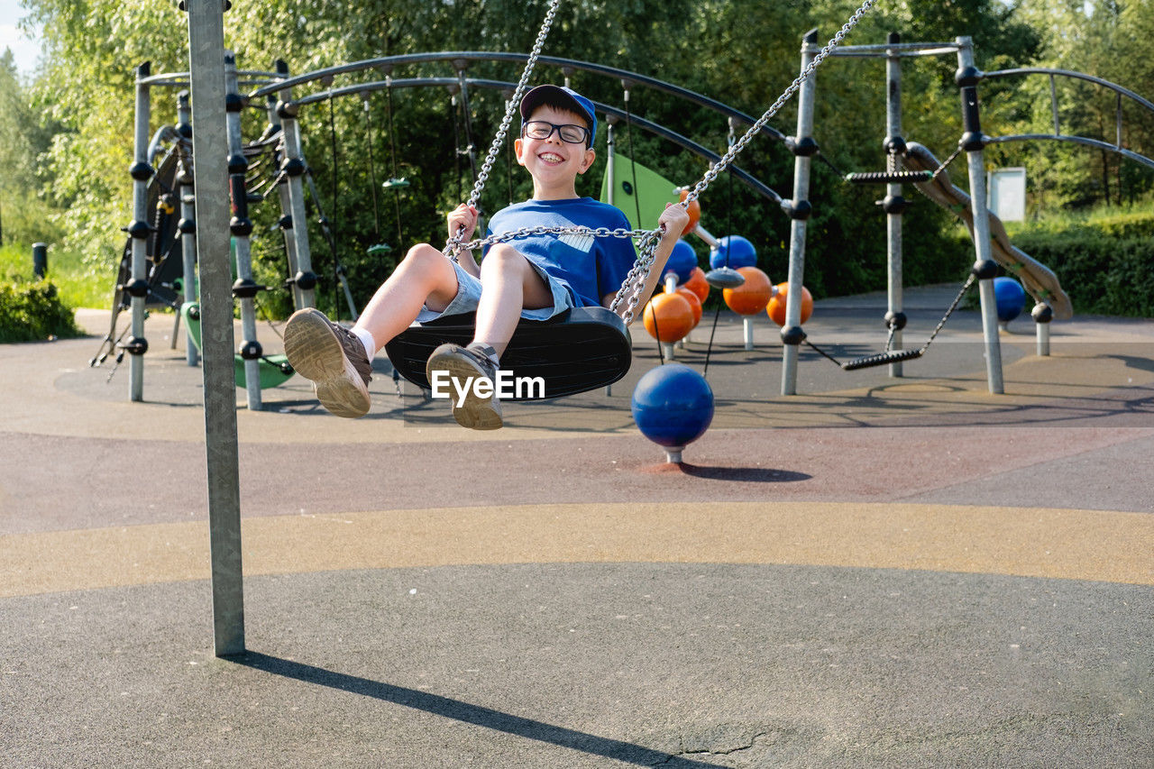 An 11 year old boy wearing glasses and a blue t-shirt is riding a swing in a park on a playground