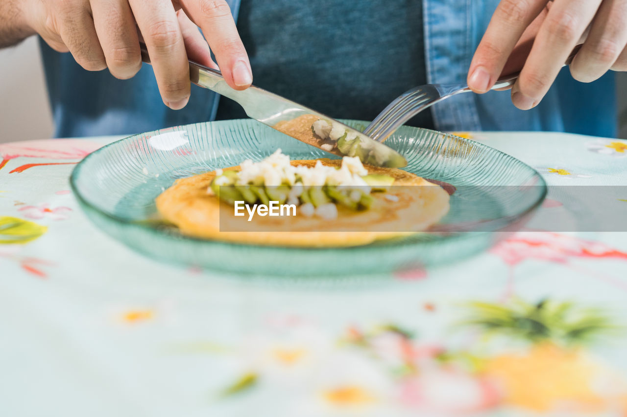 A plate of pancakes with cheese and avocado is on the table. an anonymous man is eating with a colorful tablecloth