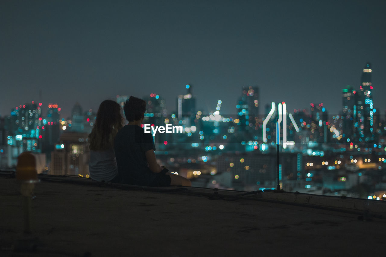 Rear view of young couple looking at cityscape while sitting on building terrace against sky