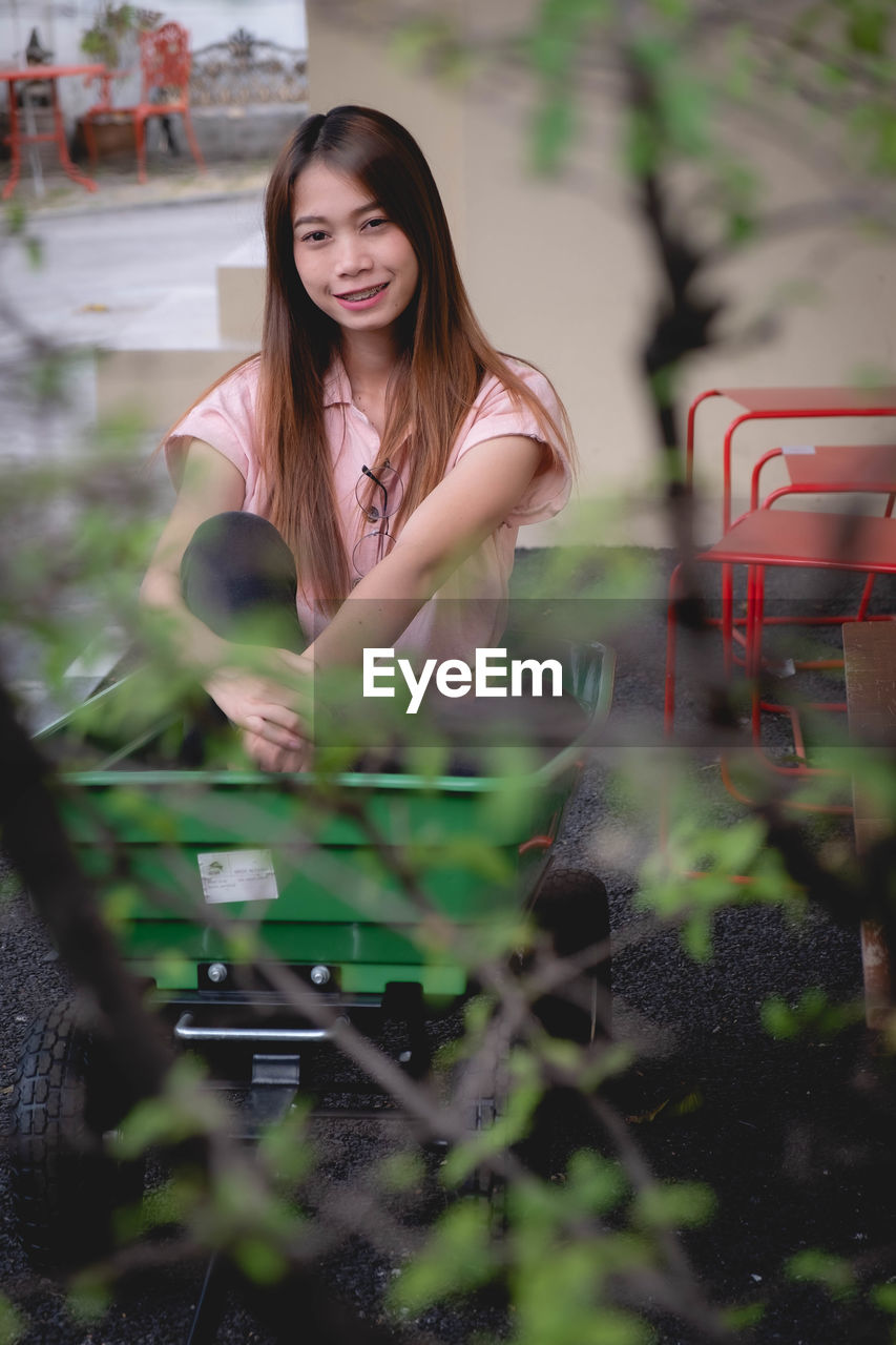 Portrait of young woman sitting in wheelbarrow against wall