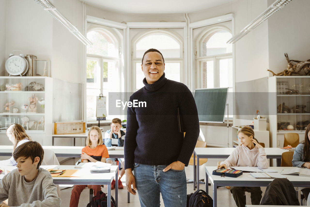 Portrait of happy male teacher with hand in pocket standing amidst students in classroom