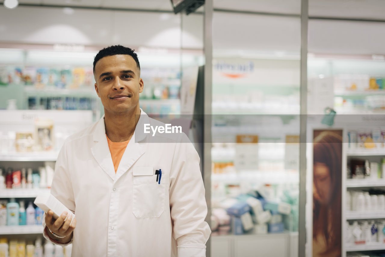 Portrait of confident owner wearing lab coat standing with medicine in pharmacy store