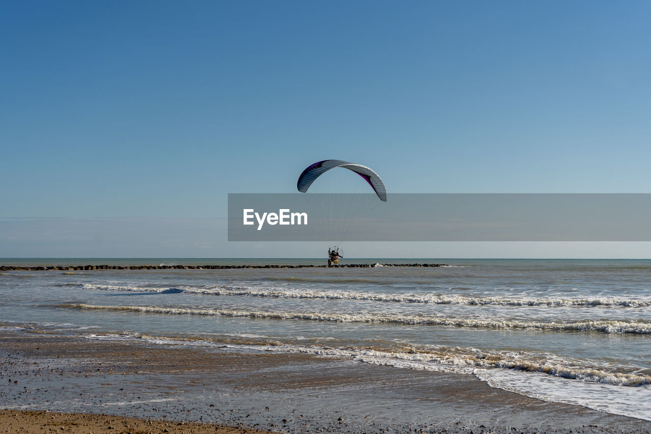 sky, sea, water, land, beach, sports, adventure, extreme sports, paragliding, nature, horizon, horizon over water, leisure activity, motion, scenics - nature, parachute, beauty in nature, joy, clear sky, flying, coast, sand, exhilaration, holiday, windsports, wave, ocean, environment, vacation, trip, blue, water sports, tranquility, day, mid-air, surfing, one person, lifestyles, wind wave, travel destinations, kite sports, tranquil scene, outdoors, recreation, sunny, landscape, shore, full length, activity, fun, sunlight, urban skyline, travel, summer, copy space, idyllic, transportation, wind