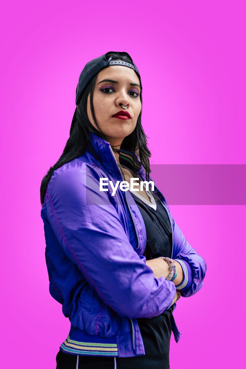 one person, purple, colored background, adult, studio shot, portrait, women, young adult, clothing, pink, standing, looking at camera, indoors, violet, photo shoot, waist up, three quarter length, blue, outerwear, pink background, fashion, hood, magenta, hairstyle, arms crossed, arts culture and entertainment, serious, person, female, jacket, looking, lifestyles, side view, purple background