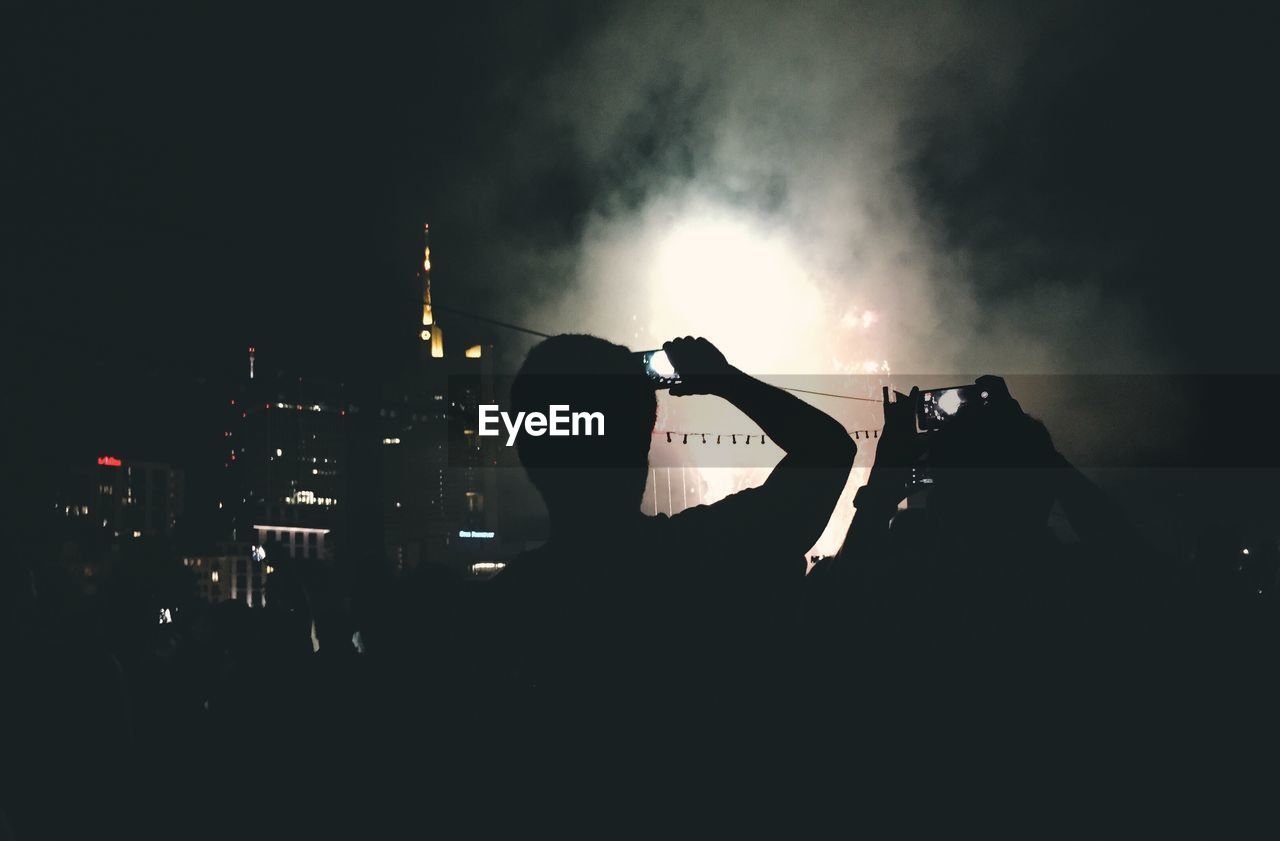 Silhouette people photographing fireworks against sky at night in city