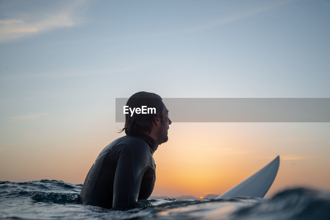 Side view of man with surfboard looking away in sea against sky during sunset