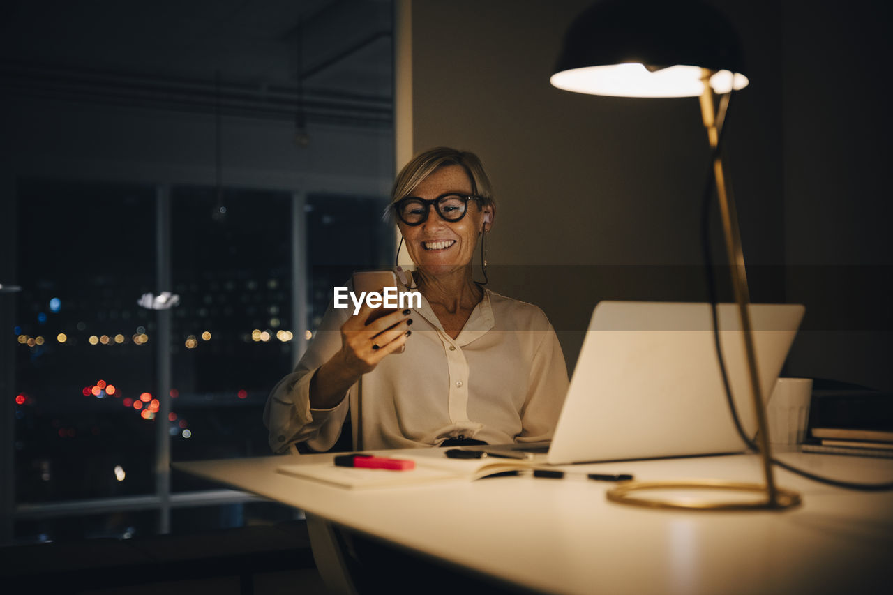 Smiling businesswoman using smart phone while sitting with laptop at illuminated desk in coworking space