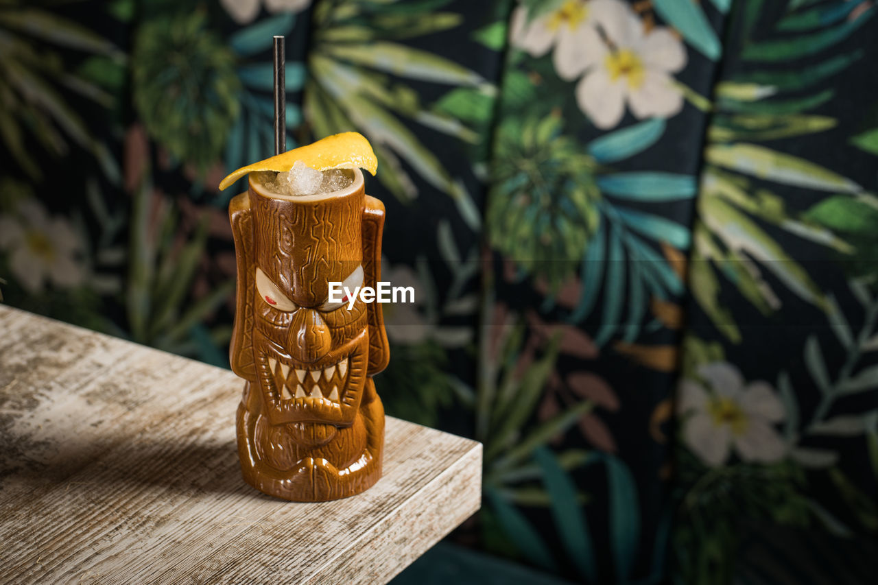 Brown sculptural tiki mug with alcohol drink decorated with straw and ice placed on table on blurred background