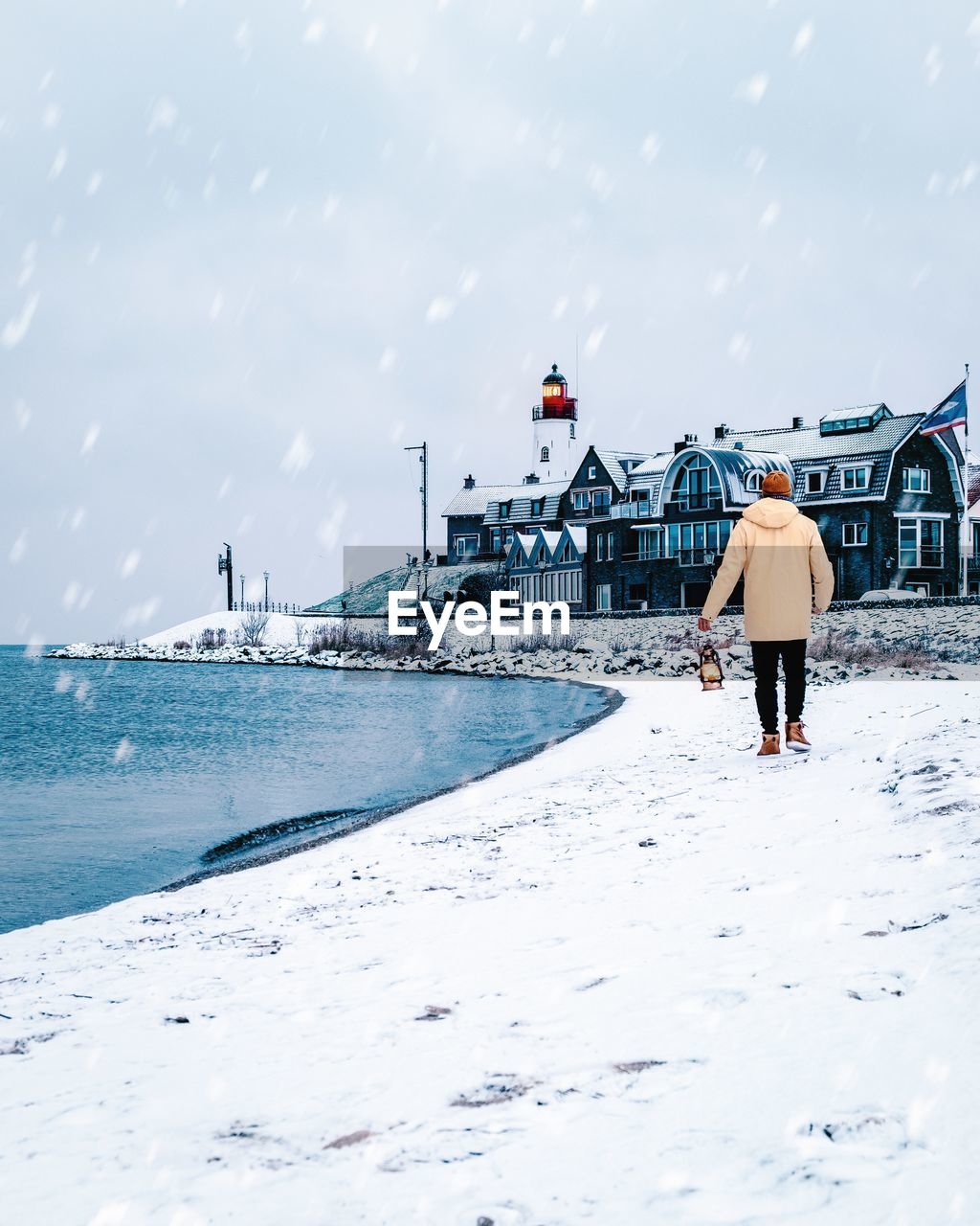 Rear view of man walking on shore against buildings during snowfall