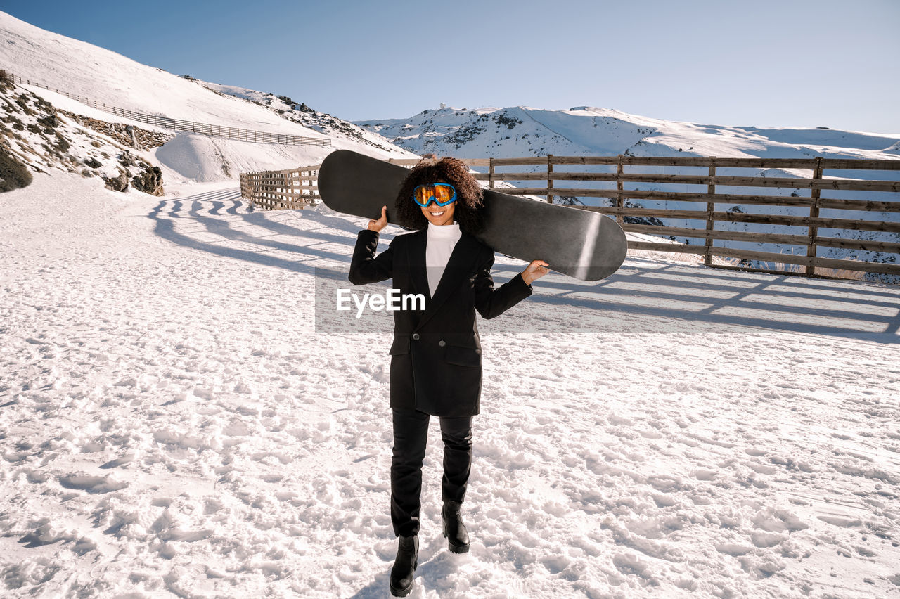 African american woman with goggles and a snowboard on a snowy mountain during winter