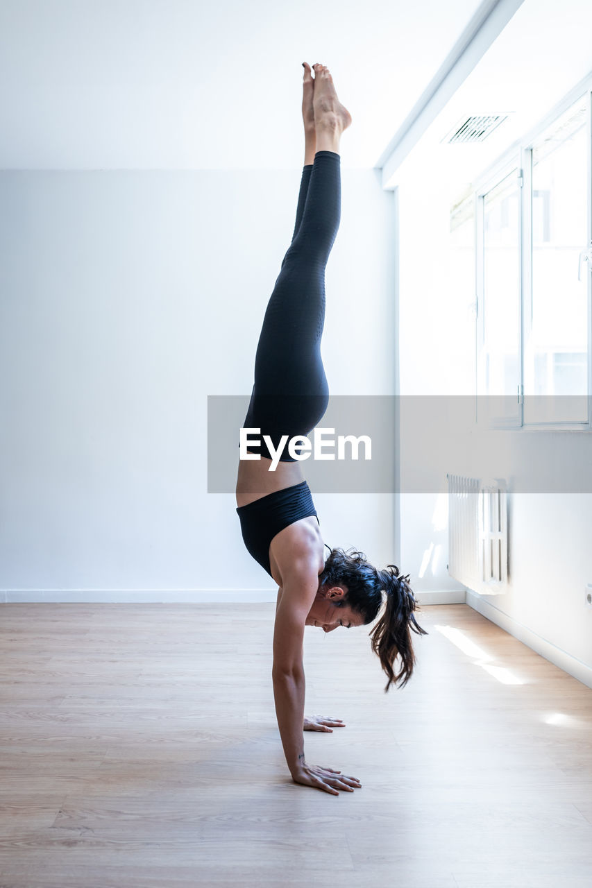 Side view of slim female athlete performing handstand on wooden floor in gym during training