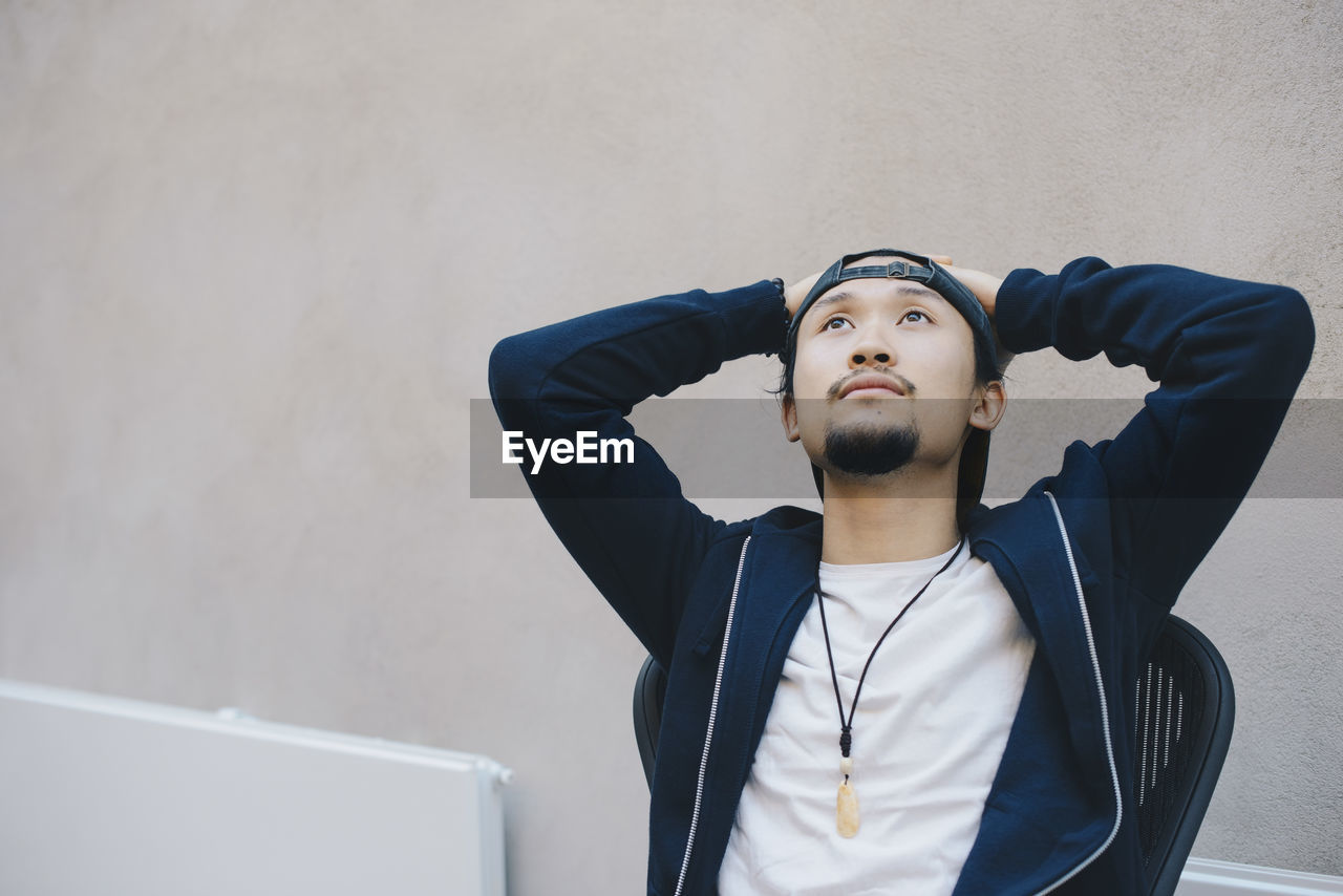 Thoughtful male computer programmer sitting with hands behind head against beige wall in office