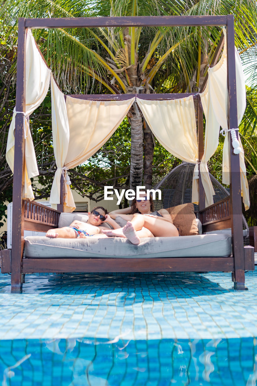 Mother and son relaxing in a bali bed near the swimming pool