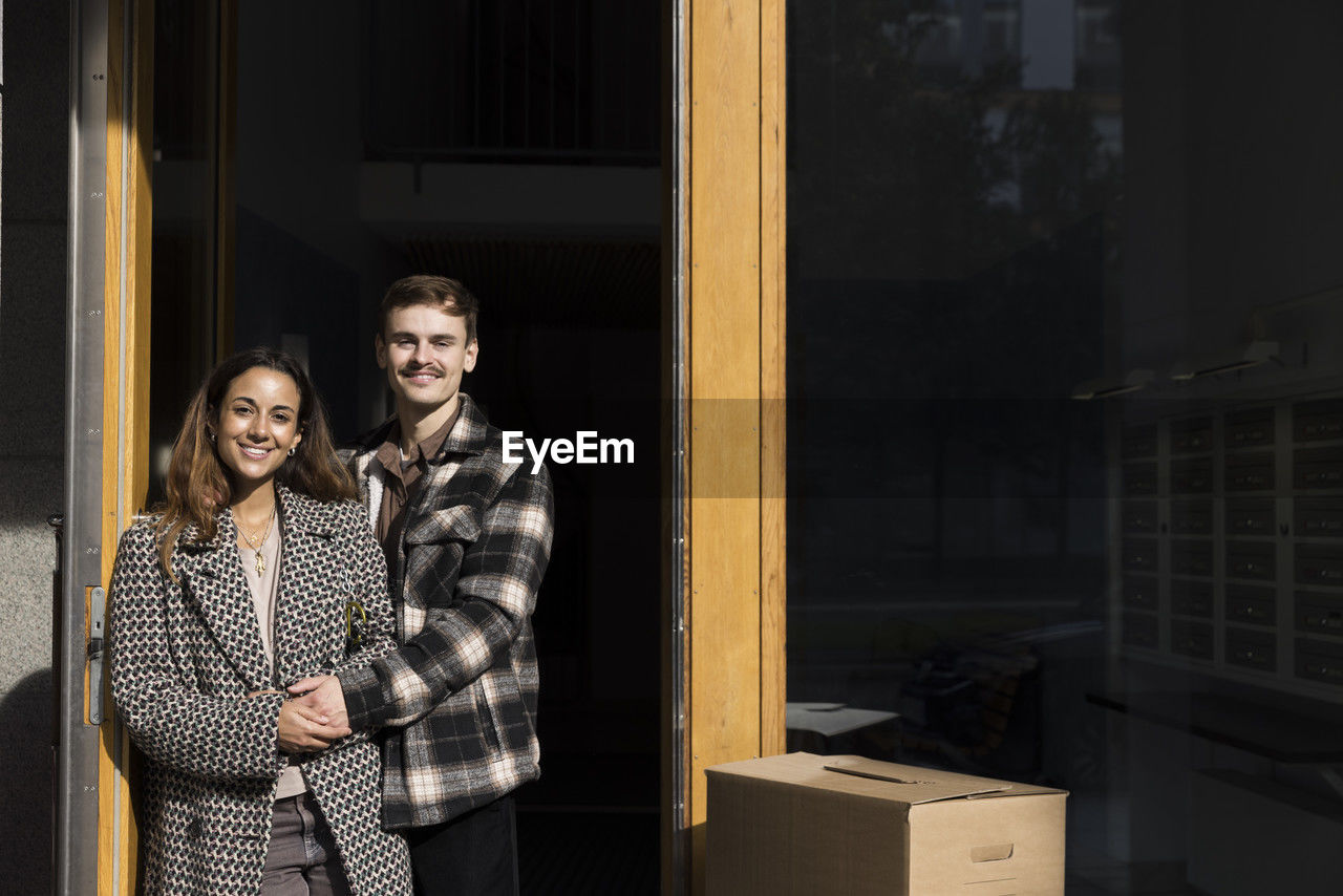 Portrait of smiling multiracial couple holding hands while standing in front of apartment