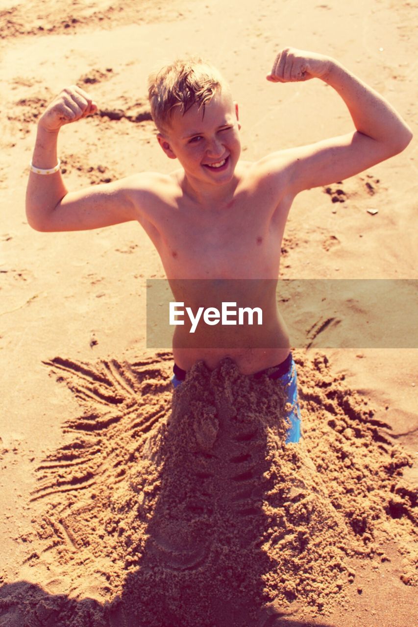 High angle portrait of smiling boy flexing muscle while standing in sand at beach