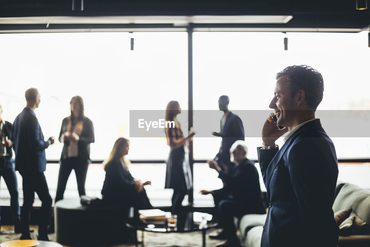 Smiling male entrepreneur talking on smart phone while colleagues discussing in background at workplace