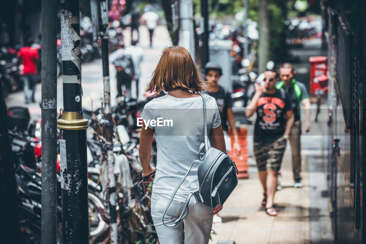 Rear view of backpack young woman walking on footpath in city