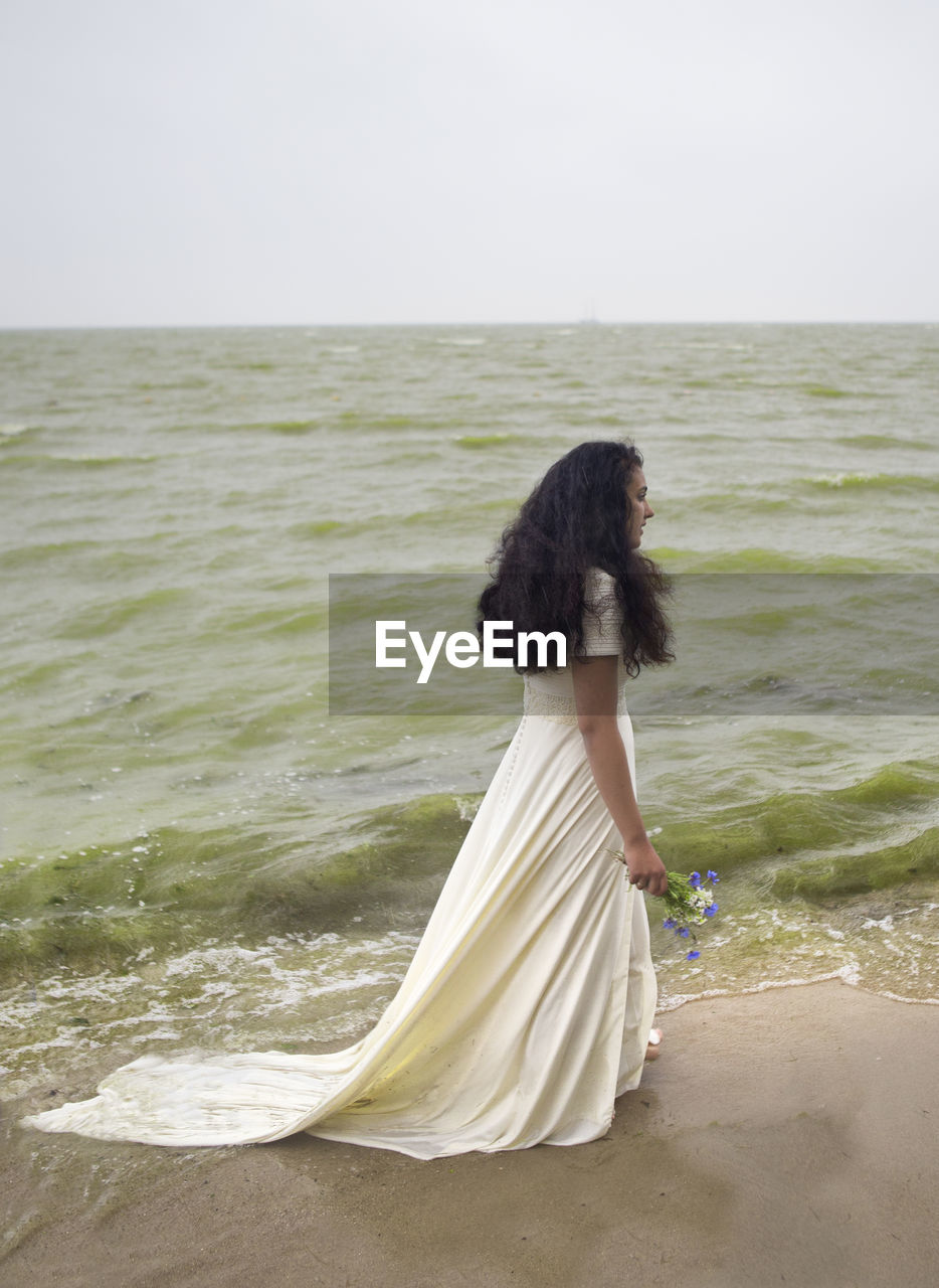 REAR VIEW OF WOMAN AT BEACH AGAINST SKY