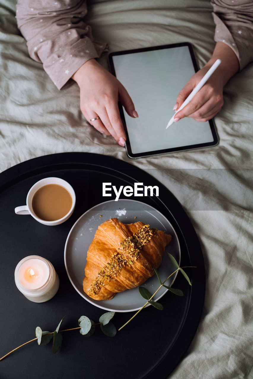 Woman using digital tablet for drawing while lying on bed and enjoying croissant with cappuccino	
