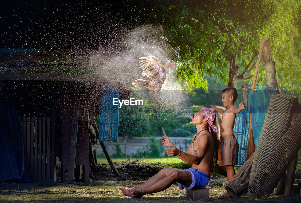 Side view of boy standing by father spraying water on hen in village