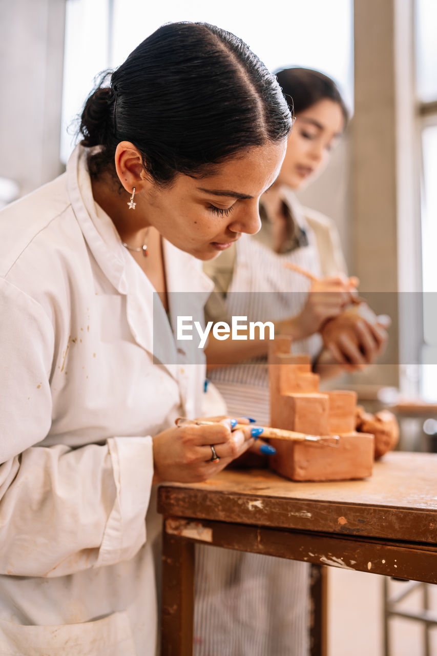 Side view merry hispanic girlfriends creating geometric shape from clay on table in spacious workshop