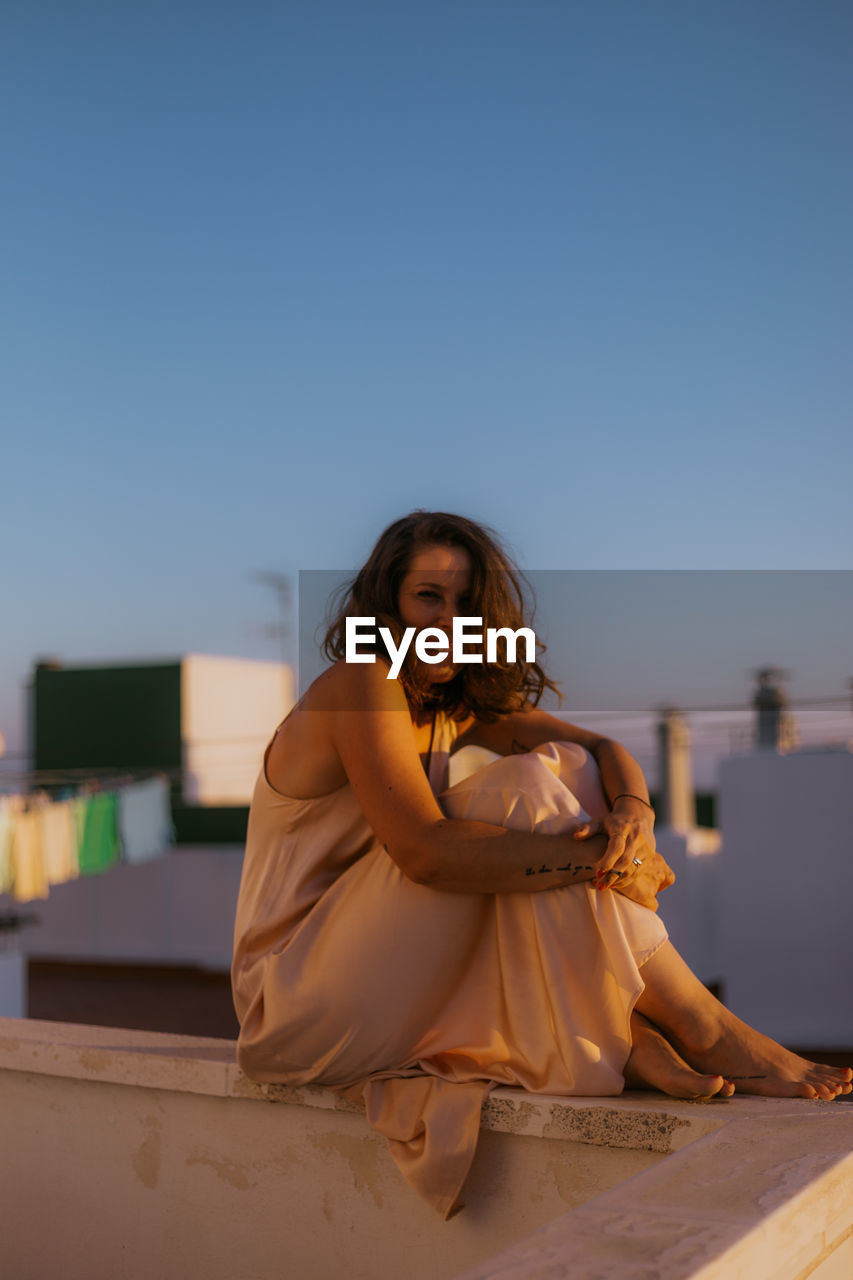 Cheerful happy woman in stylish pink silk dress sitting on fence of balcony with arms around legs and looking at camera during summer sunset