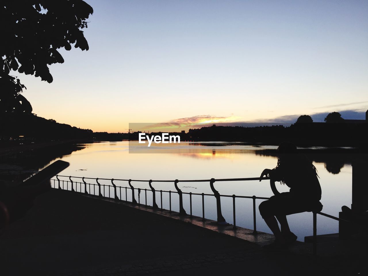 Silhouette woman sitting on railing by lake against sunset sky