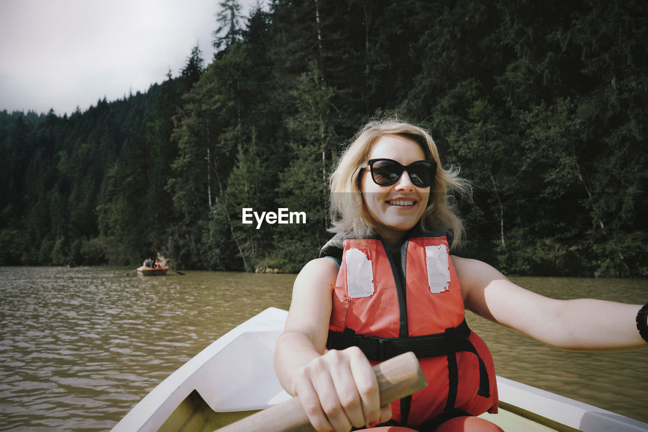Smiling woman wearing sunglasses and life jacket sitting on boat in lake